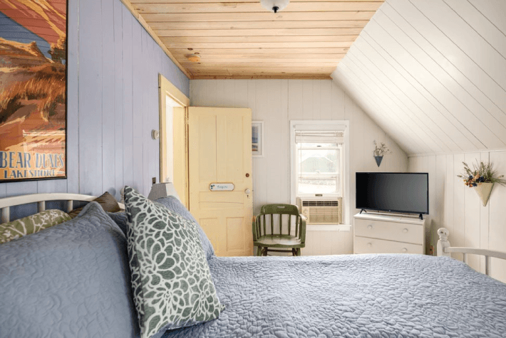 A bedroom with a blue quilted bed, a television, a small green chair, and a framed picture on the wall.