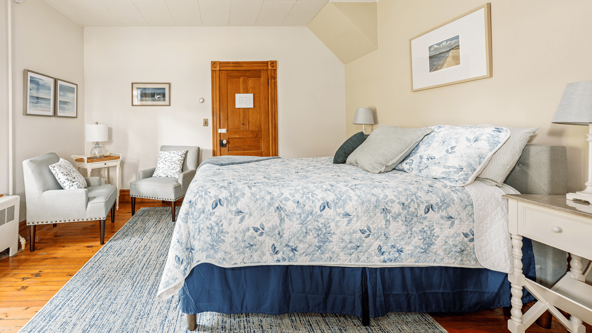 A cozy bedroom featuring a floral quilted bed, two chairs with decorative pillows, and wooden accents.