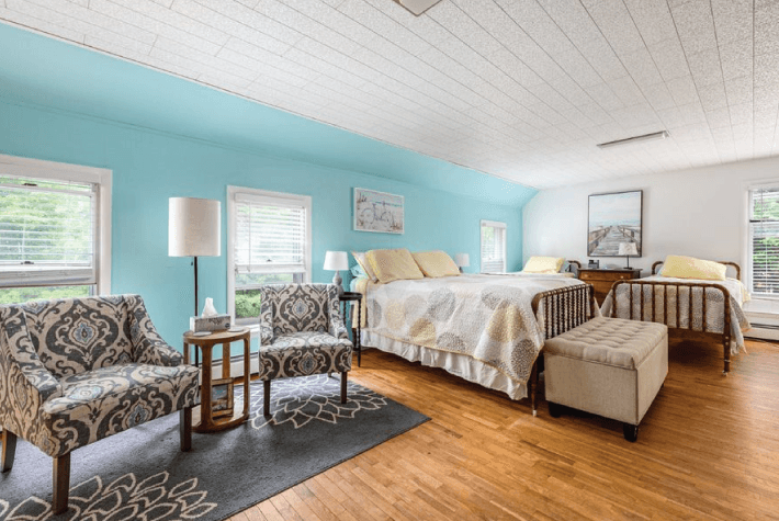 A bedroom with three beds, a blue accent wall, two patterned armchairs, and hardwood floors.