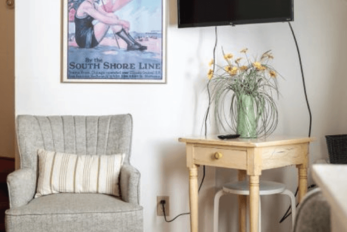 A gray armchair with a striped pillow, a light wood table with flowers, and a framed poster on the wall.