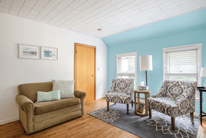 A living room with a blue accent wall, featuring a brown sofa, two patterned armchairs, and a wooden door.