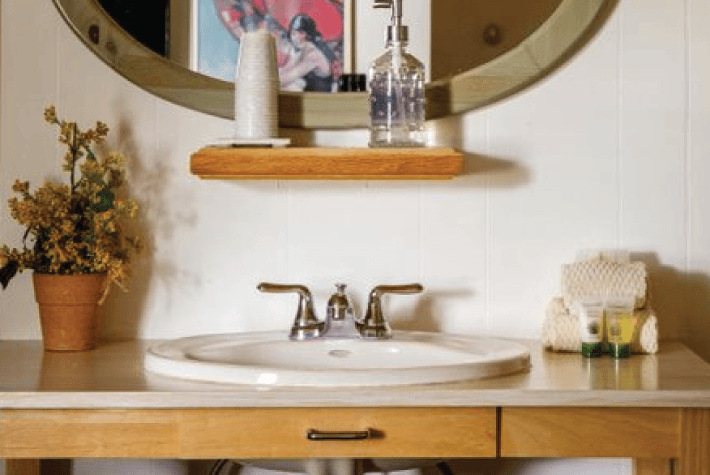 Close-up of a bathroom sink with a round mirror, wooden shelf, and various toiletries.