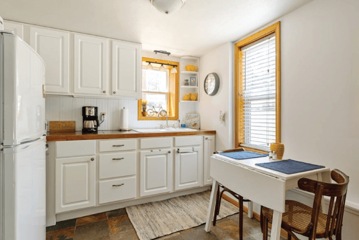 A kitchen with white cabinets, a wooden countertop, a white table, and a window with blinds.