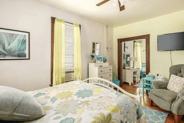 A bedroom featuring a white metal bed with a floral quilt, a gray armchair, a flat-screen TV, and a white dresser.
