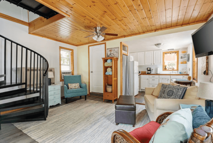 A living room with a spiral staircase, a kitchen, a teal armchair, a sofa, and a wood plank ceiling.