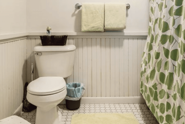 A bathroom with a white toilet, a green and white shower curtain, a towel rack, and a small basket on the toilet tank.