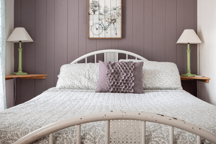 a bedroom featuring a white metal bed with a patterned quilt, two green lamps on small shelves, and a framed bicycle artwork on a dusky plum wall.