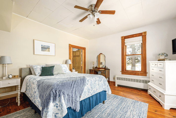 A bedroom with a blue and white bed, a white dresser, a wooden-framed window, and a ceiling fan.