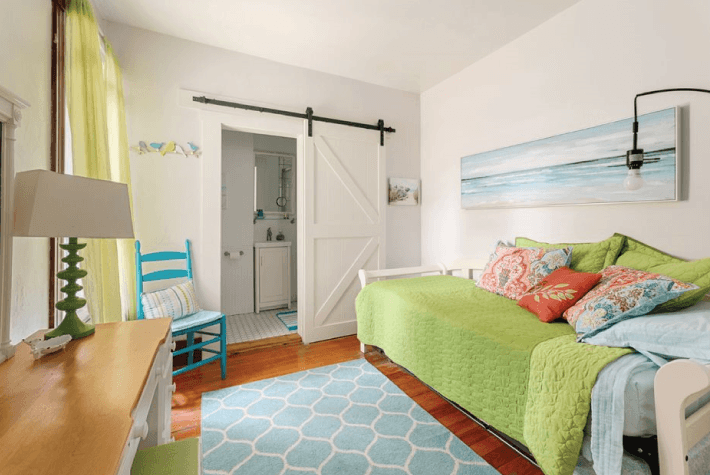 A bedroom featuring a green daybed with decorative pillows, a white sliding barn door leading to a bathroom, and a blue geometric rug.
