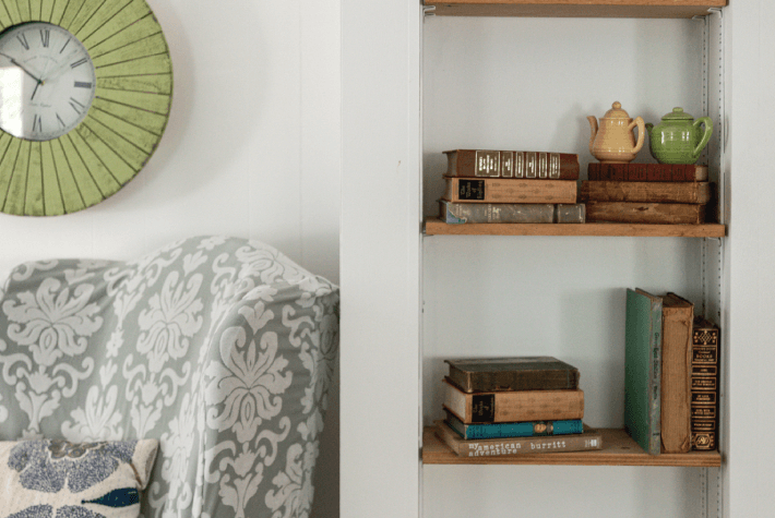 A wall with a round green clock, a patterned armchair, and built-in shelves holding books and small teapots.