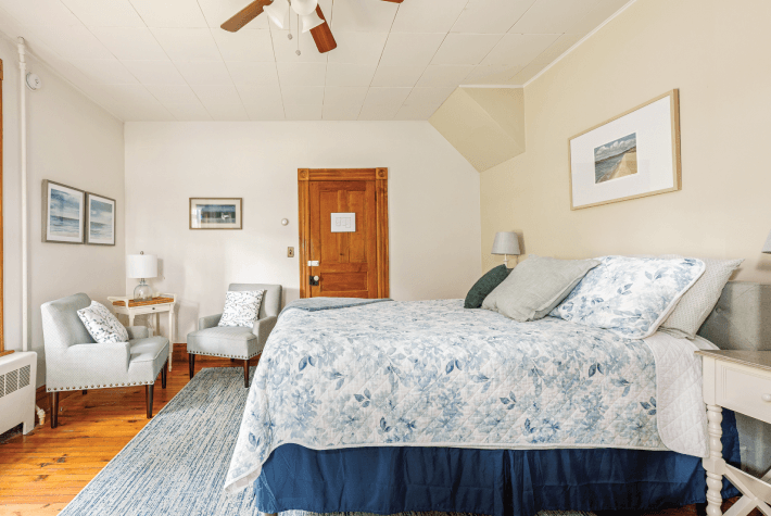 A bedroom featuring a bed with a blue and white patterned quilt, two light-colored upholstered chairs, and framed artwork.