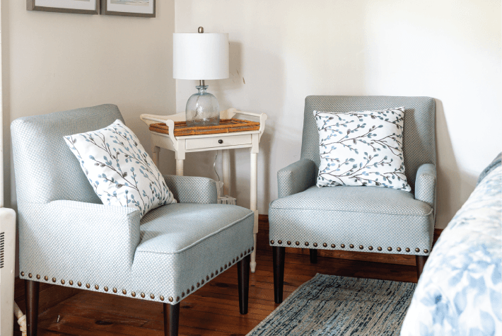 A room featuring two light blue upholstered armchairs, a small wooden side table with a lamp, and a blue rug.