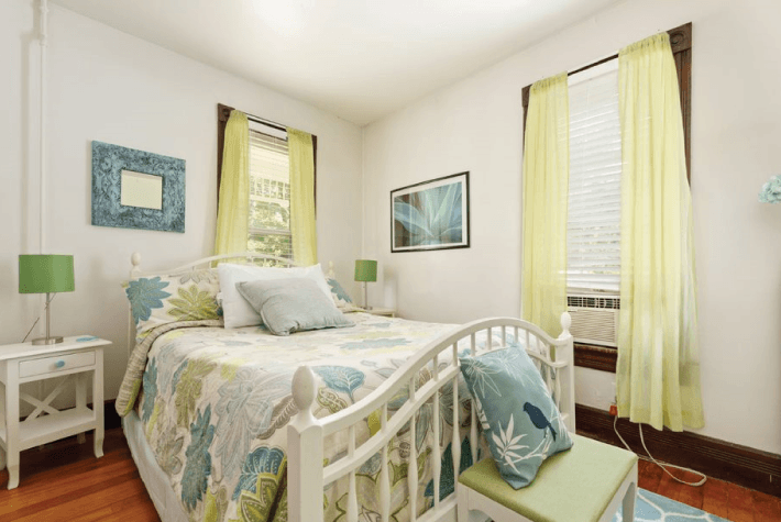A bedroom featuring a white metal bed with a floral patterned quilt, green lamps on white side tables, and yellow sheer curtains.