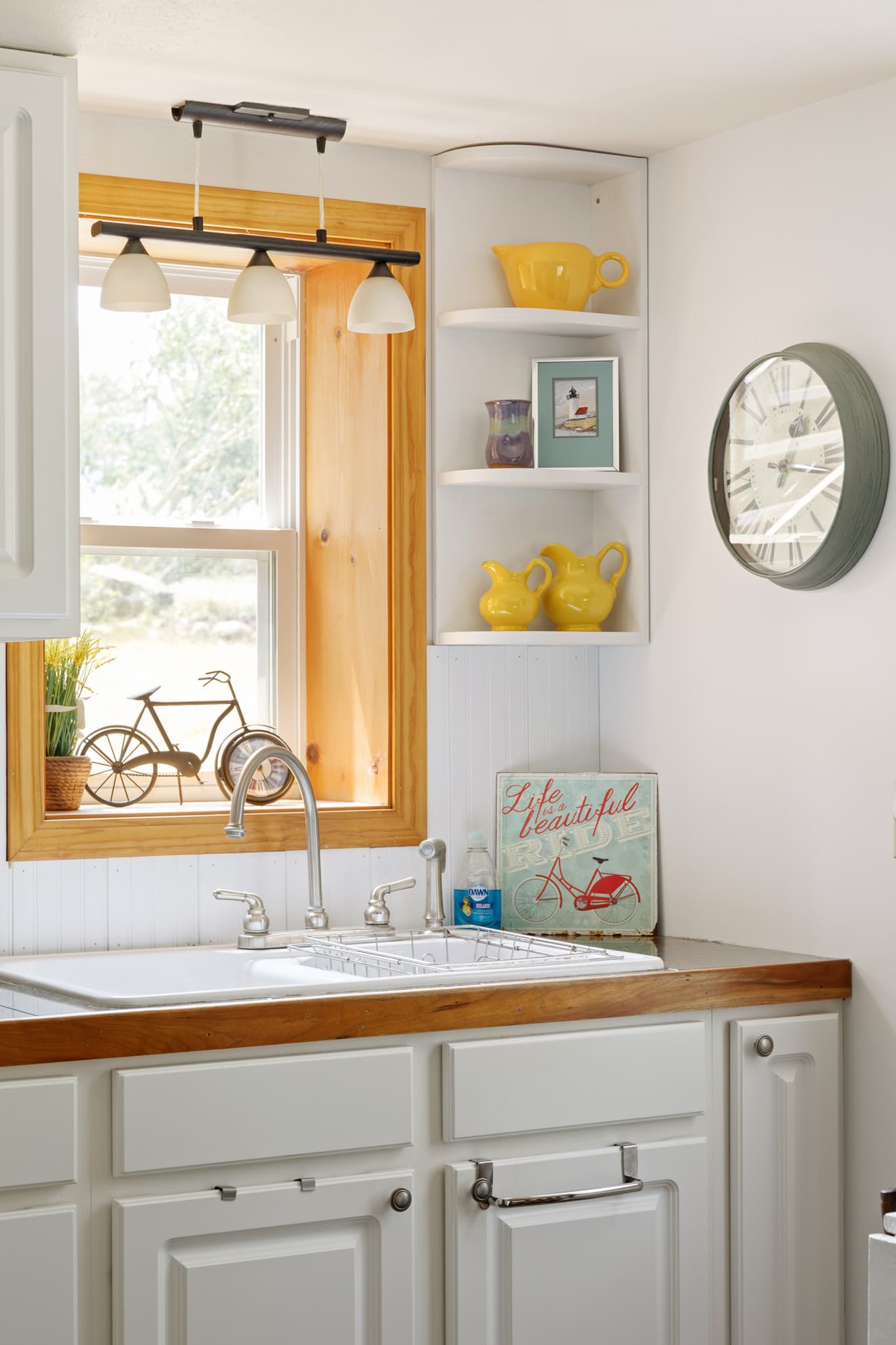 A kitchen sink with a window, a light fixture, corner shelves with yellow pitchers, and a round clock on the wall.